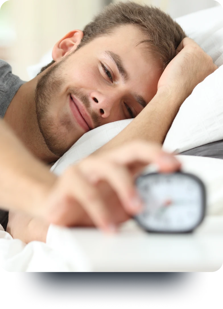 A young man, looking relaxed and lying on the bed, about to stop an alarm clock.